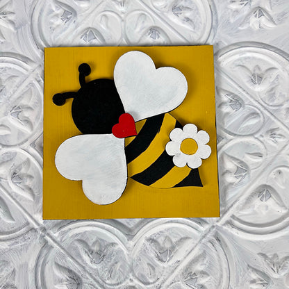 Janet's Craft Corner Bee Swappable Squares feature a handmade bee charm with white wings, black-and-yellow stripes, a red heart, and a white flower on a yellow square—perfect whimsical home decor for any textured white surface.
