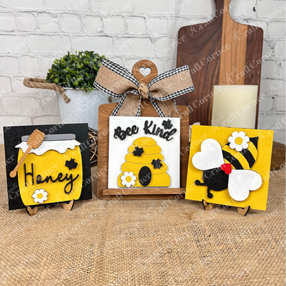 The Bee Swappable Squares DIY kit by Janet's Craft Corner includes three designs—a bee, a honey jar labeled "Honey," and a beehive arranged with farmhouse accents on a burlap surface with a white brick wall behind them for charming home decor.