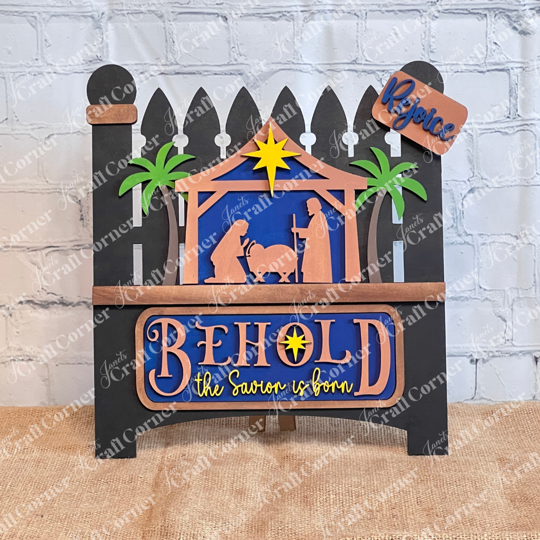 The Garden Fence with Insert by Janet's Craft Corner features a nativity scene silhouette of Mary, Joseph, and baby Jesus in a stable with palm trees and a star. It includes the text "Behold the Savior is born" on a brick wall backdrop and comes as an interchangeable home decor kit.