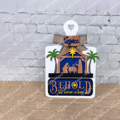 Janet's Craft Corner presents a DIY Breadboard Interchangeable Base with an insert featuring a nativity scene of Mary, Joseph, and baby Jesus under a star. The sign reads "BEHOLD the Savior is born," with "Rejoice" at the top, adorned with a bow and festive ornaments for warmth and celebration.