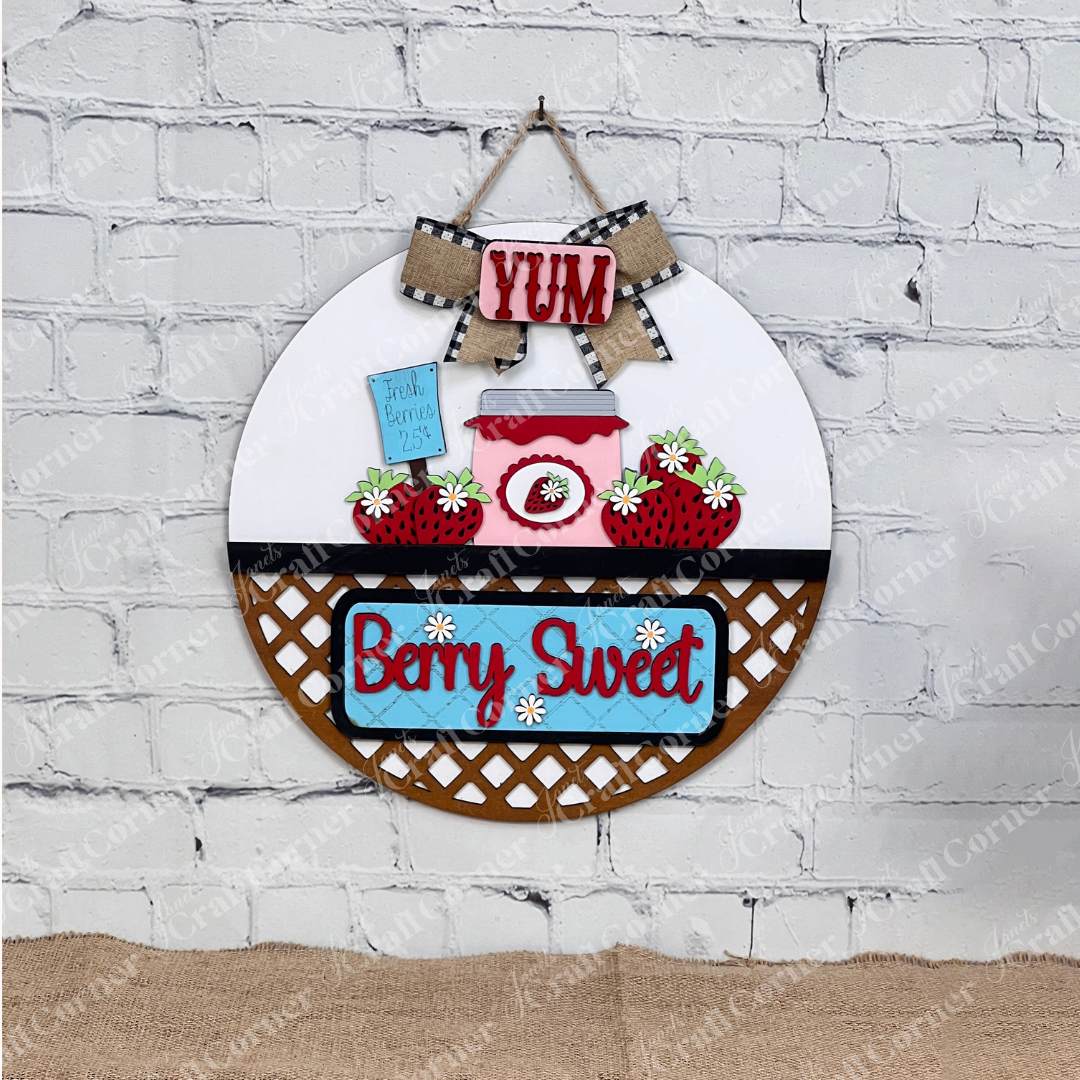 The Berry Sweet Interchangeable Insert by Janet's Craft Corner features a round decor piece with "Berry Sweet," strawberries, a jam jar, and "YUM" on a wooden basket-like background, perfect for adding charm to farmhouse décor when hung on a white brick wall.