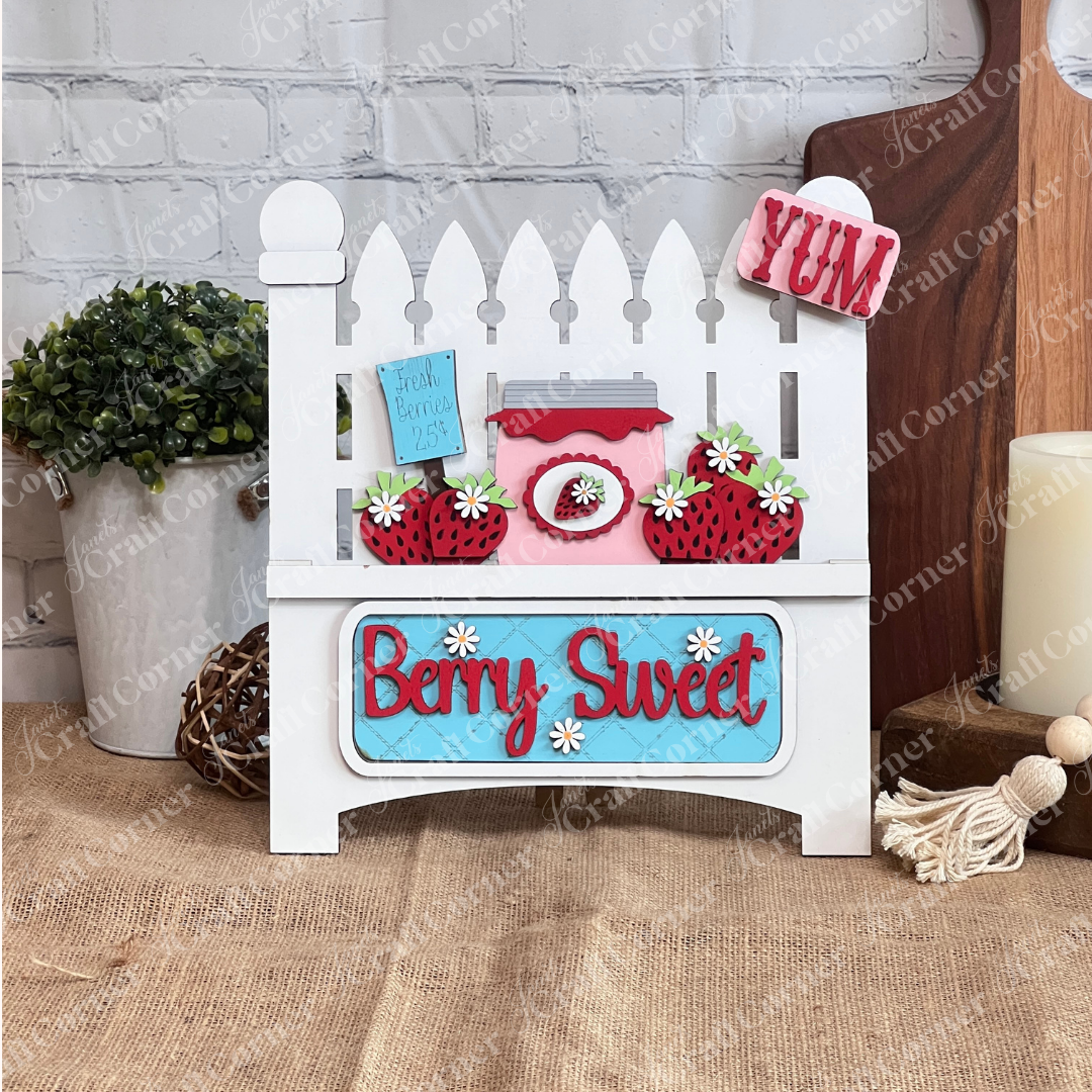 The Berry Sweet Interchangeable Insert by Janet's Craft Corner is a DIY seasonal decor kit featuring a picket fence design, jar of jam, strawberries, flowers, and signs "Berry Sweet," "Fresh Berries 25¢," and "YUM." It sits against a white brick wall on a burlap surface.