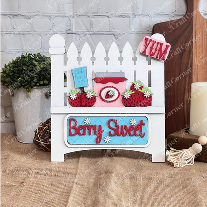 The Berry Sweet Interchangeable Insert by Janet's Craft Corner is a DIY seasonal decor kit featuring a picket fence design, jar of jam, strawberries, flowers, and signs "Berry Sweet," "Fresh Berries 25¢," and "YUM." It sits against a white brick wall on a burlap surface.