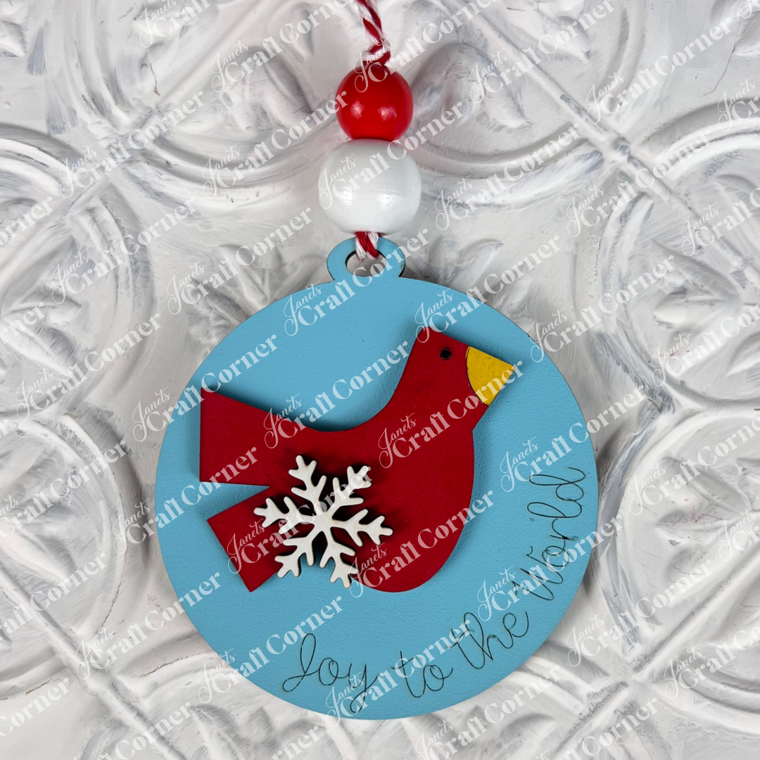 A round Christmas ornament from Janet's Craft Corner's DIY Decoration Kits features a charming light blue design with a red bird and white snowflake decoration. The cursive text "Joy to the World" enhances its appeal. It hangs gracefully from a red and white string, set against an embossed white background—perfect for adding to your DIY kit collection.