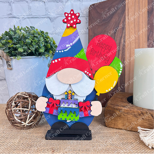 A vibrant Birthday Gnome from Janet's Craft Corner holds a cake and balloons, one of which is inscribed with "Happy Birthday." It features a hat topped with a star. Perfect as home décor, it stands on a textured surface surrounded by plants, candles, and charming wall decorations.