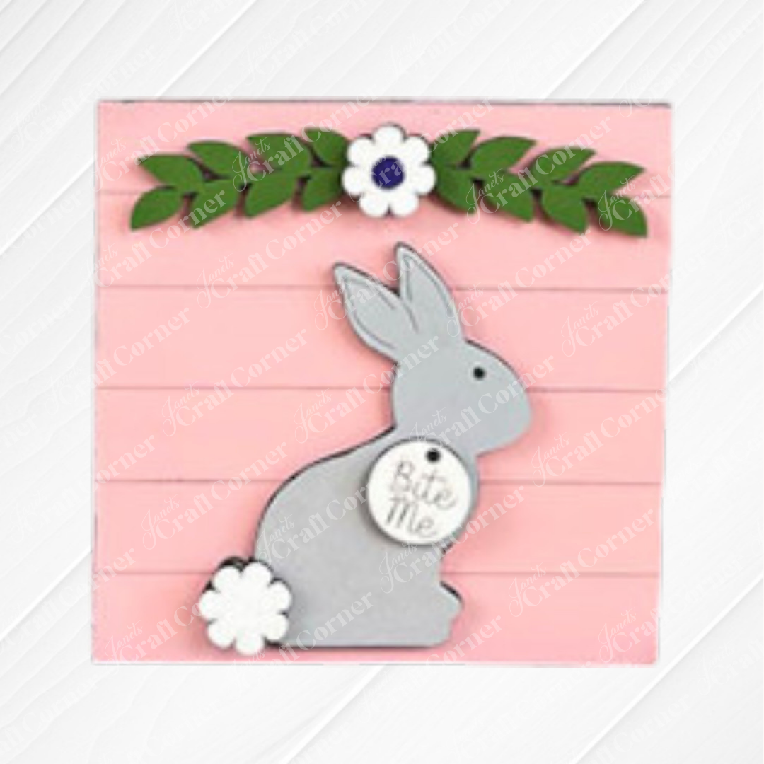 The Easter Bunny Squares by Janet's Craft Corner feature a gray bunny with a white flower near its tail, sitting on a pink background with green leaves and white flowers. A round "Bite Me" tag adds charm—perfect for tiered tray decor.
