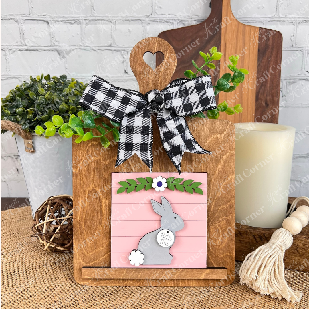The Easter Bunny Squares by Janet's Craft Corner features a wooden stand with a heart cutout holding a pink sign with a gray bunny, "Bite Me" text, green leaves, and white flower—ideal rustic tiered tray decor for Swappable Square Displays.