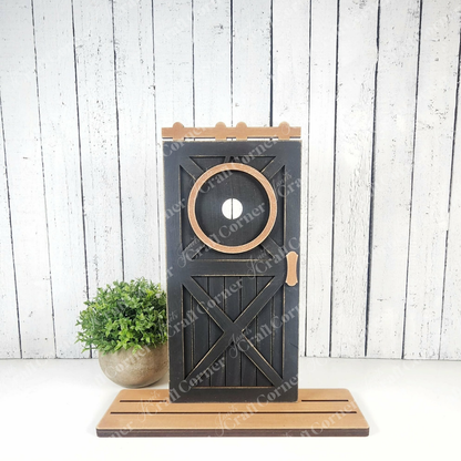 Janet's Craft Corner Farmhouse Porch Interchangeable Base is a DIY seasonal home decor kit featuring a mini black barn door with wood accents, perfect as wall art or a rustic accent against white wooden planks. Set of 1; plant not included.