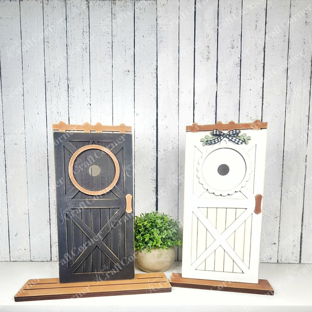 Janet’s Craft Corner Farmhouse Porch Interchangeable Base DIY kit features two black and white farmhouse-style door accents with wooden handles and a potted plant, perfect for seasonal wall decor. Set of 1.
