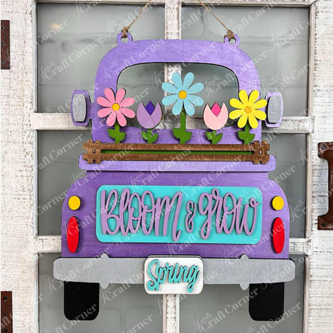 A charming DIY home decor kit from Janet's Craft Corner features an antique-style purple truck crafted from wood, adorned with vibrant flowers in its bed. The rear of the truck is embellished with "Bloom & Grow" in teal, while a small sign below displays "Spring." This versatile piece hangs elegantly on a white door and invites creativity with its interchangeable inserts.