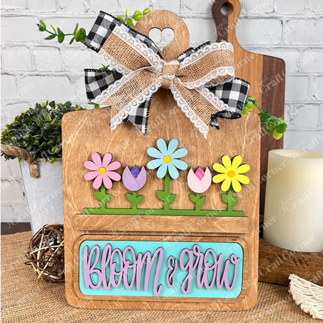 The Breadboard Interchangeable Base with Insert by Janet's Craft Corner is ideal for creative DIYers. Featuring colorful flower cutouts and "Bloom & Grow" in pink and blue, it includes a plaid and lace bow, complemented by items like plants, candles, and cutting boards against a brick wall.
