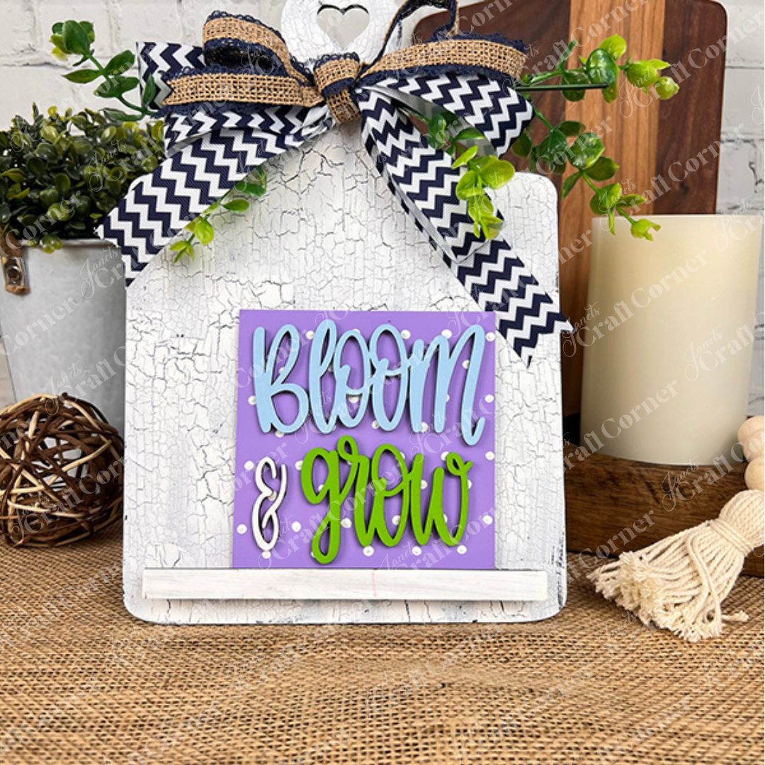 Janet's Craft Corner's Bloom & Grow Squares is a decorative white sign with a black and white chevron bow and greenery, featuring playful “Bloom & Grow” lettering—perfect for tiered trays or rustic wooden table displays.