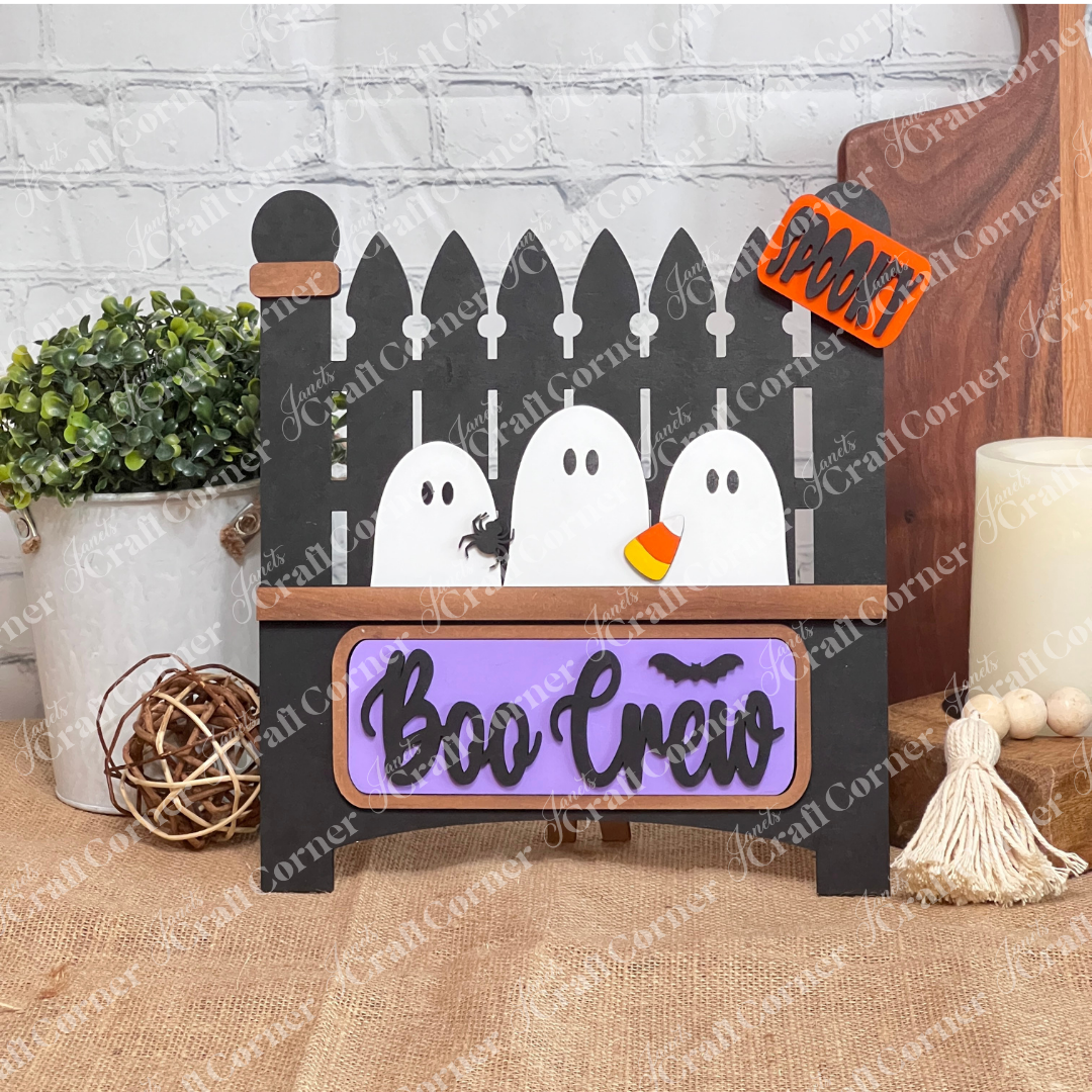Janet's Craft Corner offers the Boo Crew Interchangeable Insert DIY kit, featuring a sign with three white ghosts behind a black fence, embellished with candy corn and spider accents. The purple plaque reads "Boo Crew" with a bat, and has an attached "spooky sidekick" sign.