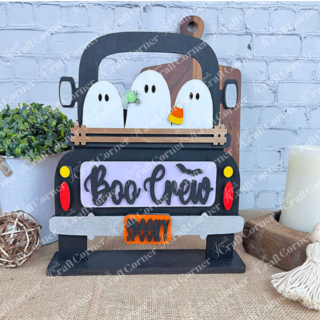 A decorative display showcases a charming trio of ghost figures in the back of a black truck, crafted with Janet's Craft Corner's Boo Crew Interchangeable Insert DIY home decor kit. The truck's sign displays "Boo Crew" with a bat graphic and features a "Spooky" license plate, all set against a backdrop of white brick decorated with plants and other decor elements.