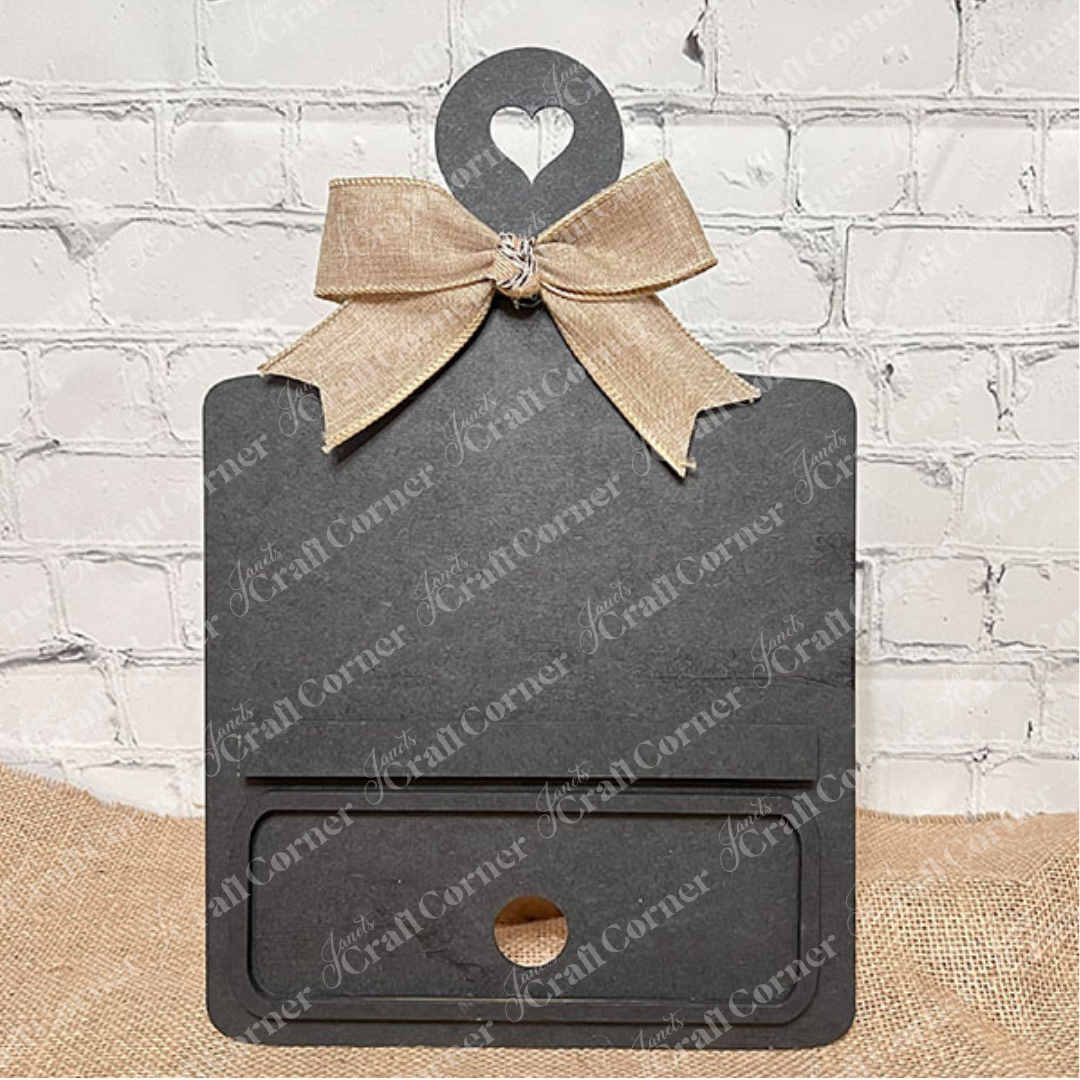 Janet's Craft Corner offers the Breadboard Classic Interchangeable Base with Insert DIY kit. This decorative black board has a heart handle, gingham bow, minimalistic face markings, and interchangeable accessories—ideal for DIY enthusiasts against a burlap backdrop.