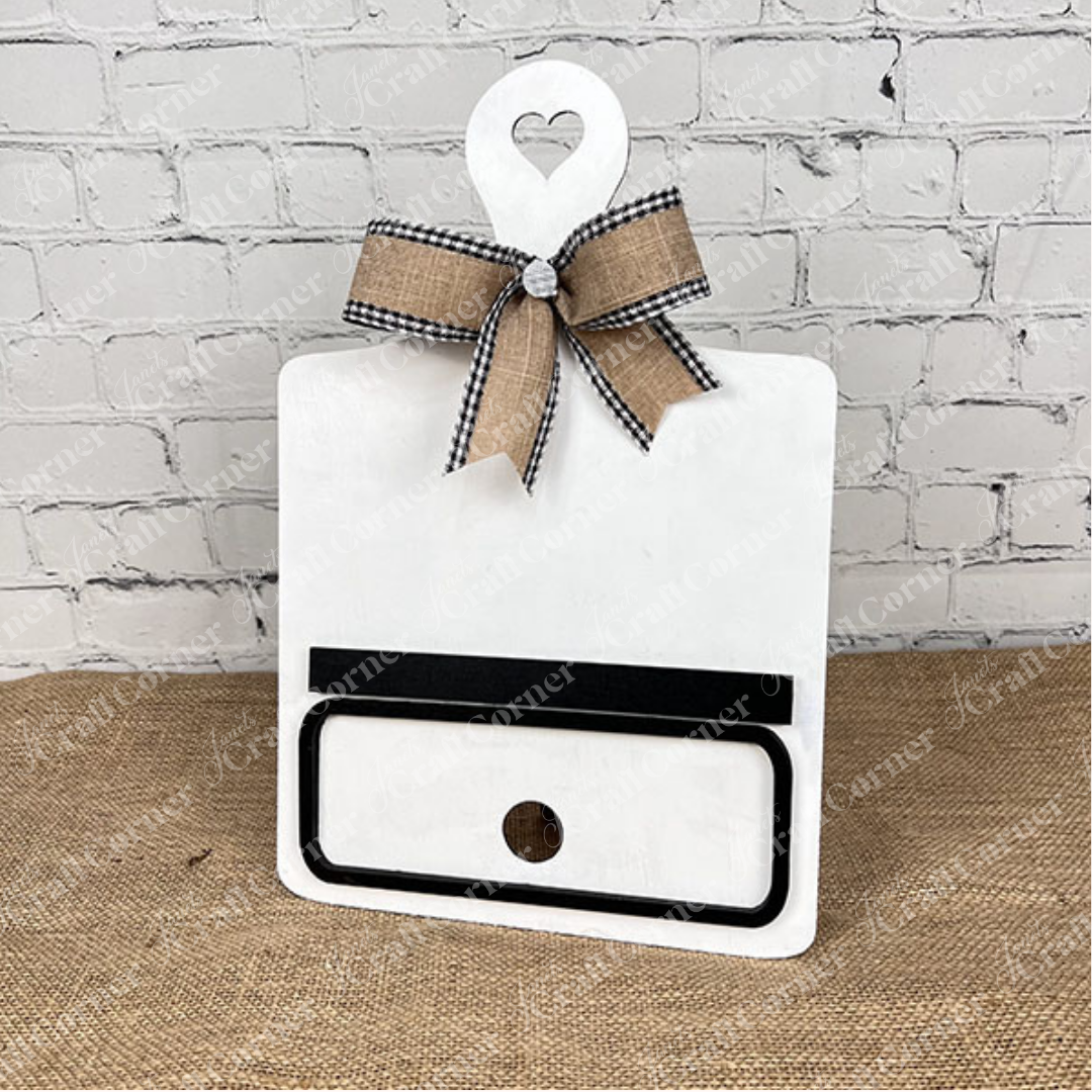 Janet's Craft Corner offers the Breadboard Classic Interchangeable Base with Insert DIY kit. This decorative white board has a heart handle, gingham bow, minimalistic face markings, and interchangeable accessories—ideal for DIY enthusiasts against a burlap backdrop.