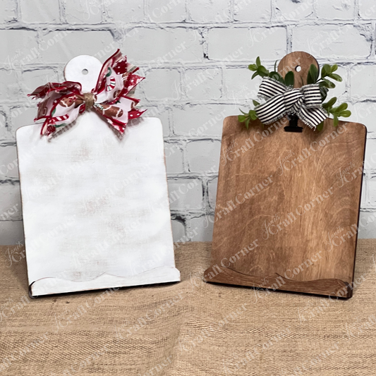 Two Breadboard Sign Holders from Janet's Craft Corner, one in a plain brown finish and the other whitewashed, each featuring leafy embellishments and a striped bow at the top, are displayed against a rustic white wooden backdrop. Perfect for farmhouse decor enthusiasts or as part of your DIY home decor craft kit.