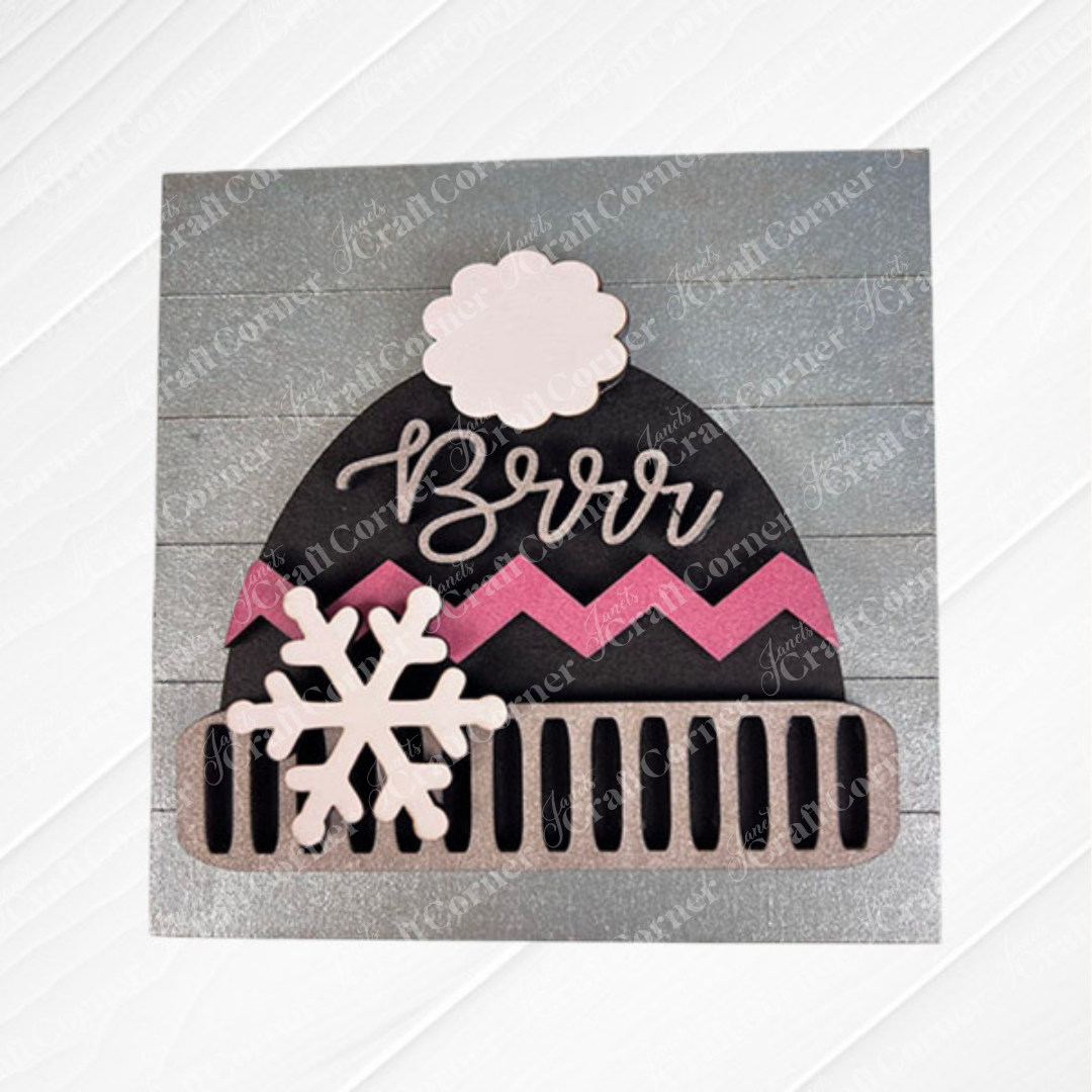 The "Hello Winter Squares" sign by Janet's Craft Corner showcases a winter hat with a white pom-pom, pink zigzags, snowflakes, and "Brrr" on the front atop a gray wood-textured background—perfect for seasonal home decor.