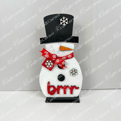 Janet’s Craft Corner Snowman Shelf Sitters feature a snowman with a black top hat, red scarf, carrot nose, and snowflake accents. “Brrr” in red letters makes it perfect winter décor for any festive home.