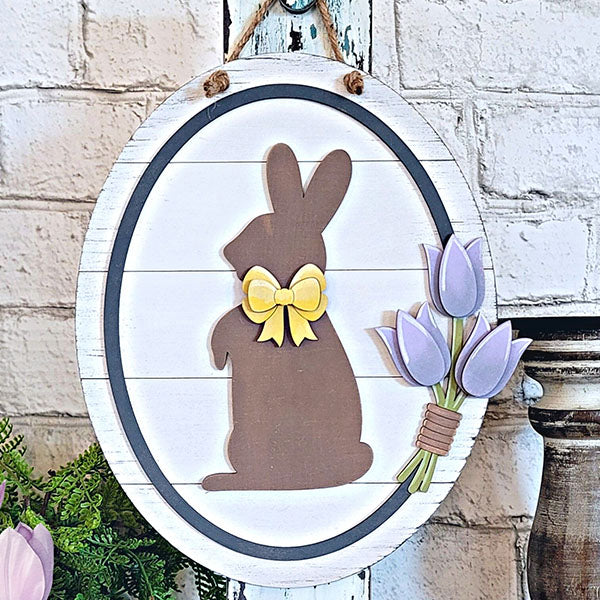 The Bunny and Tulips Sign from Janet's Craft Corner is an oval wooden plaque ideal for Easter decoration. It showcases a bunny silhouette with a yellow bow, accompanied by three purple tulips on the right, and hangs elegantly against a white brick wall.