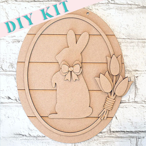 Easter decor DIY Kit: The Bunny and Tulips Sign from Janet's Craft Corner is a delightful woodland set featuring an oval frame with a bunny and tulips design. The bunny, accessorized with a bow, sits next to three tulips against a white brick wall backdrop. Ideal for crafting enthusiasts wanting to infuse their space with festive charm.