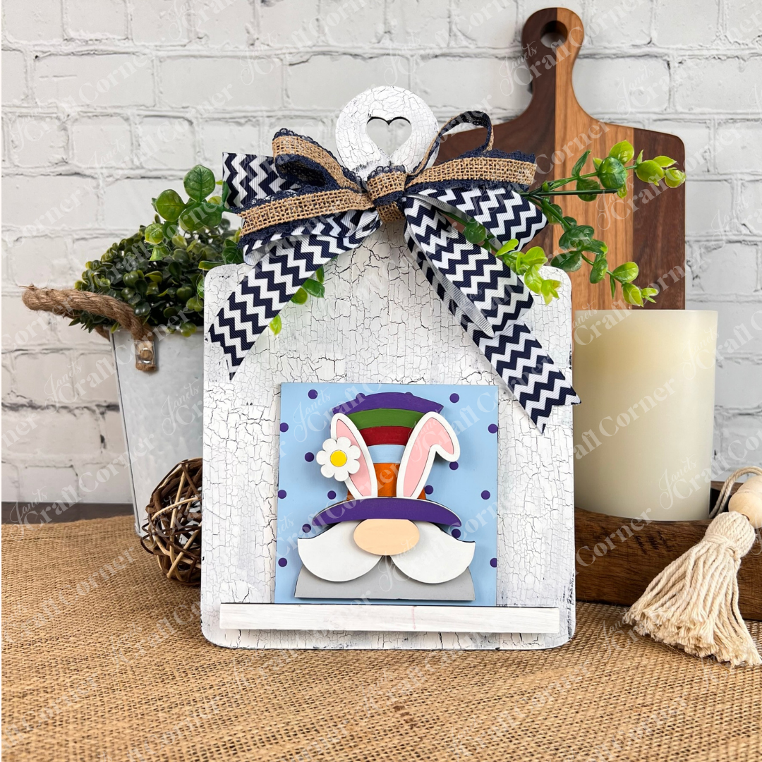 A whimsical bunny-face sign with a colorful hat and chevron bow stands on rustic white, accented with greenery—perfect to pair with Janet's Craft Corner Hello Easter Squares for charming farmhouse decor.