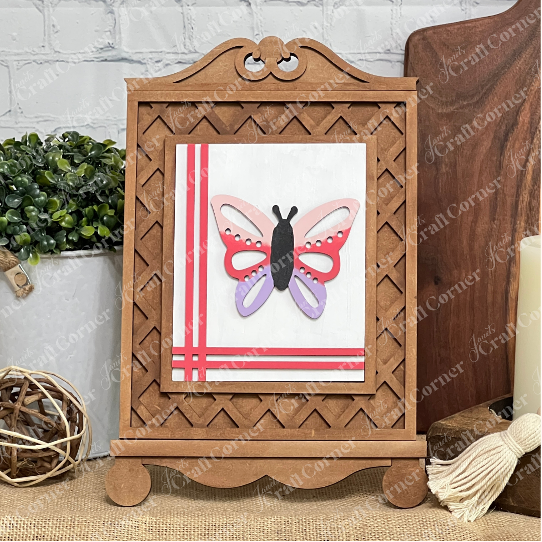 Janet's Craft Corner Cottage Frame for Switchable Farmhouse Signs (set of 1) is displayed with a handmade butterfly card on an unfinished wood easel, accented by decorative plants and a candle against a white brick background.