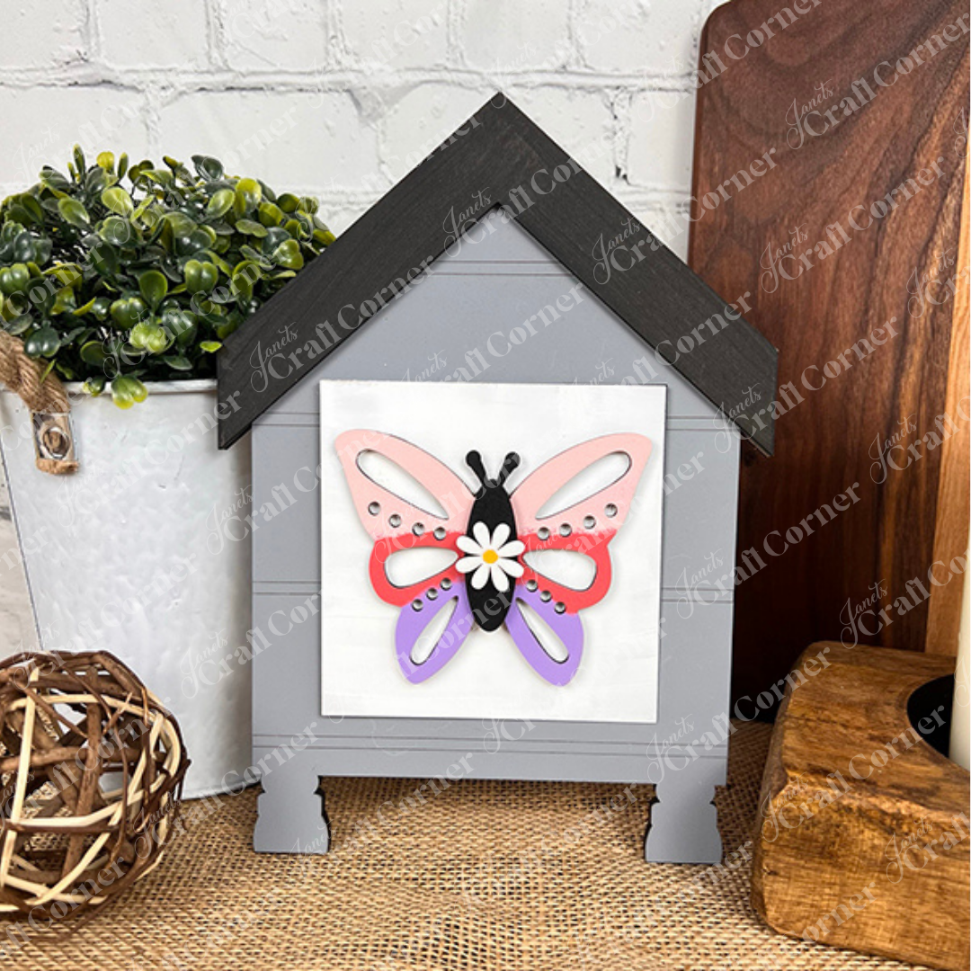 The Hello Spring Squares by Janet's Craft Corner is a gray house-shaped box with a butterfly and white flower design, perfect for tiered tray decor. Pair with other Swappable Squares wood cut-outs for a charming spring display.
