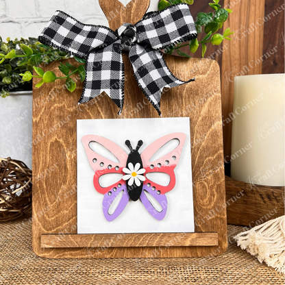Janet's Craft Corner "Hello Spring Squares" features a colorful butterfly wood cut-out with pink, red, and purple wings, a daisy center, black-and-white plaid bow, greenery accents, and wooden stand—perfect tiered tray decor.