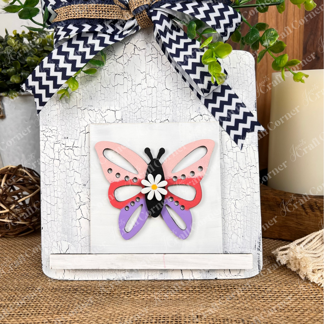 Janet's Craft Corner’s Hello Spring Squares sign features a colorful butterfly with pink, red, black, and purple wings, a white flower accent, distressed white background, chevron-patterned bow, and greenery—ideal tiered tray decor.