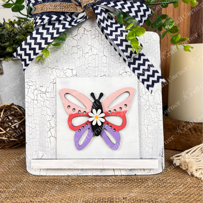 Janet's Craft Corner’s Hello Spring Squares sign features a colorful butterfly with pink, red, black, and purple wings, a white flower accent, distressed white background, chevron-patterned bow, and greenery—ideal tiered tray decor.