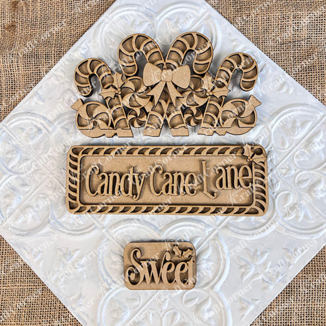 Displayed on a textured background are three wooden craft pieces from Janet's Craft Corner: a hand-painted DIY home decor kit featuring candy canes with a bow, an interchangeable insert titled "Candy Cane Lane," and a smaller piece embellished with the word "Sweet" and decorative leaves.