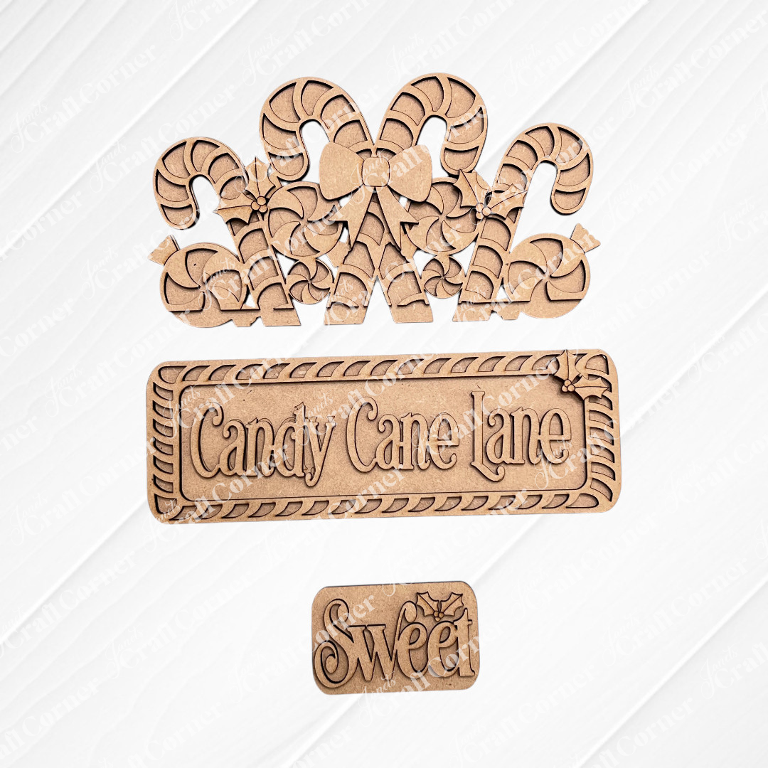 Displayed on a textured background are three wooden craft pieces from Janet's Craft Corner: a hand-painted DIY home decor kit featuring candy canes with a bow, an interchangeable insert titled "Candy Cane Lane," and a smaller piece embellished with the word "Sweet" and decorative leaves.
