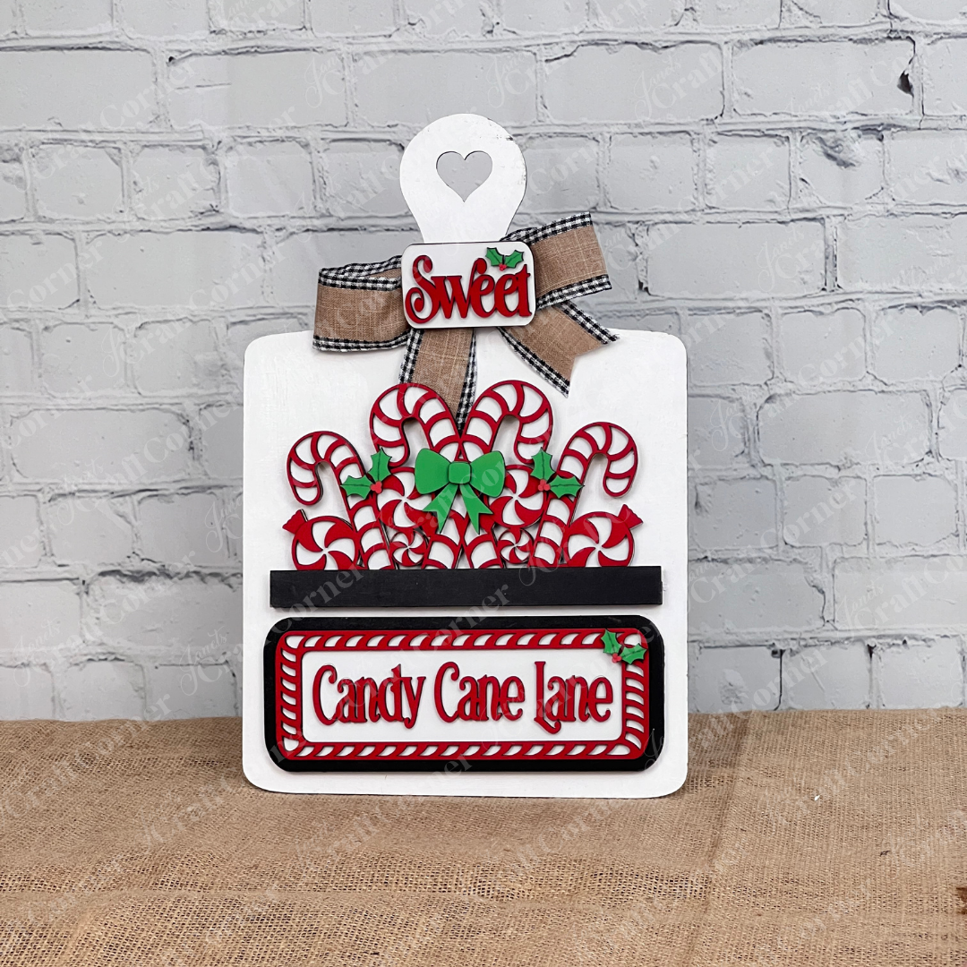 Displayed against a white brick wall is a decorative white breadboard adorned with candy canes and green bows, showcasing Janet's Craft Corner's hand-painted DIY home decor kit, the "Candy Cane Lane Interchangeable Insert," featuring signs that read "Candy Cane Lane" and "Sweet.