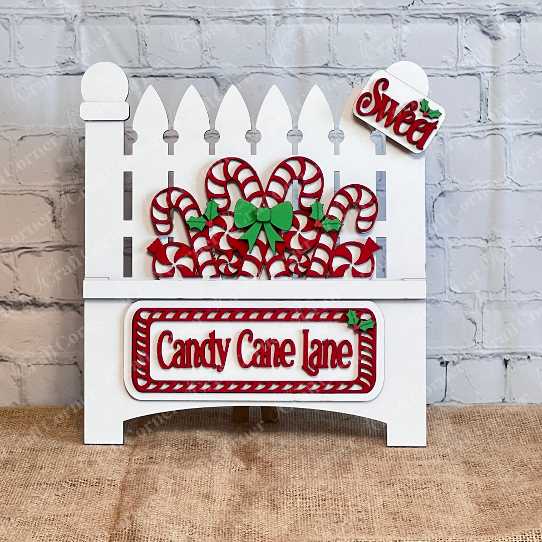 Janet's Craft Corner offers a "Garden Fence with Insert" DIY decor kit. This set includes a festive white picket fence decoration featuring interchangeable inserts like red and white candy canes and green holly, labeled "Candy Cane Lane" in red letters with a small "Sweet" sign on top, against a charming textured wall background.