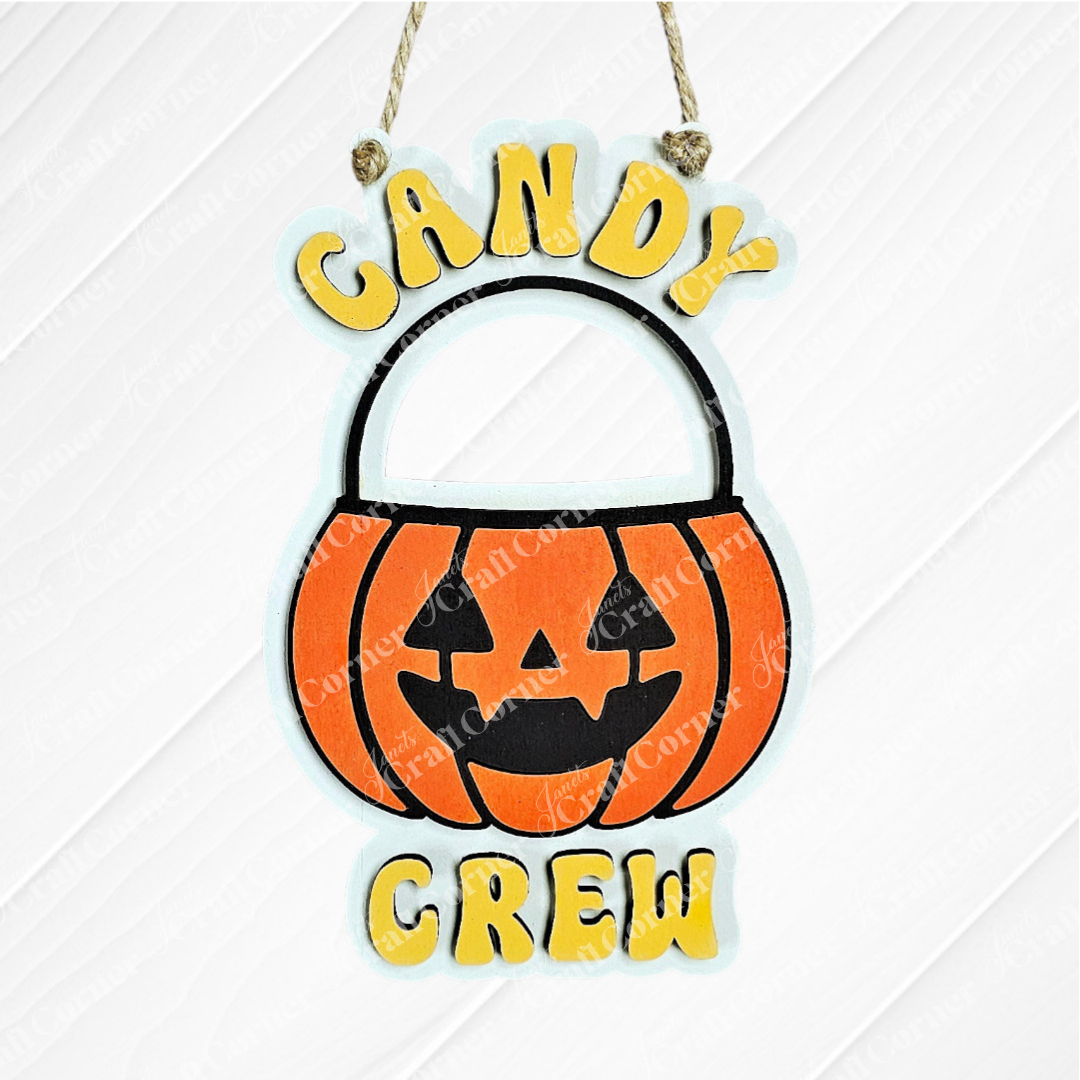 The Candy Crew Kids Pop-Out by Janet's Craft Corner features a smiling jack-o'-lantern candy bucket and "CANDY CREW" in yellow, hanging by rope—a perfect Halloween decor piece to add festive charm to your home.