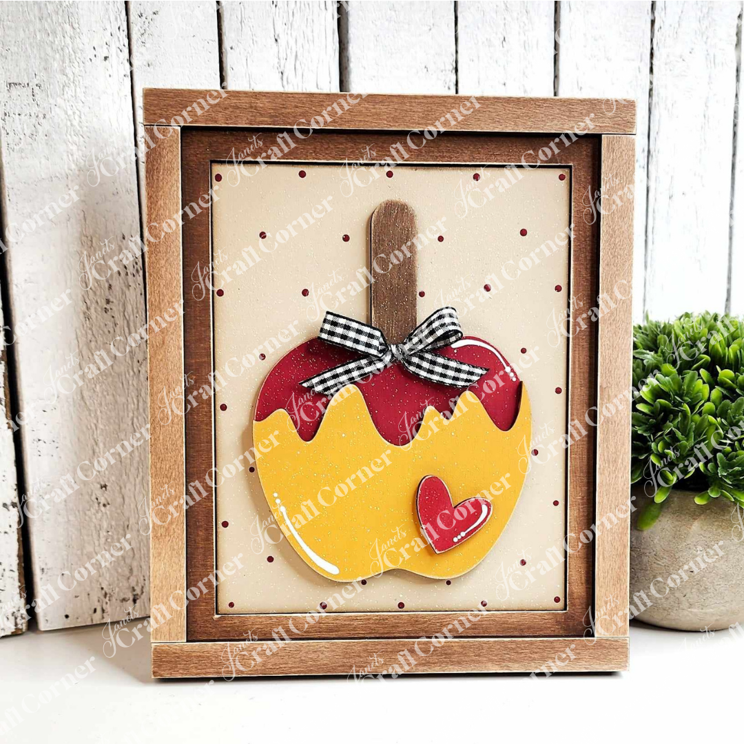 This Caramel Apple Interchangeable Sign, part of the DIY home decor craft kit by Janet's Craft Corner, features a framed design of a yellow apple with a red-dipped top and heart detail. It is elegantly adorned with a black and white checkered bow on the wooden stem. Displayed against a dotted backdrop, this sign stands on a wooden base accompanied by a small plant, making it ideal for interchangeable home décor.
