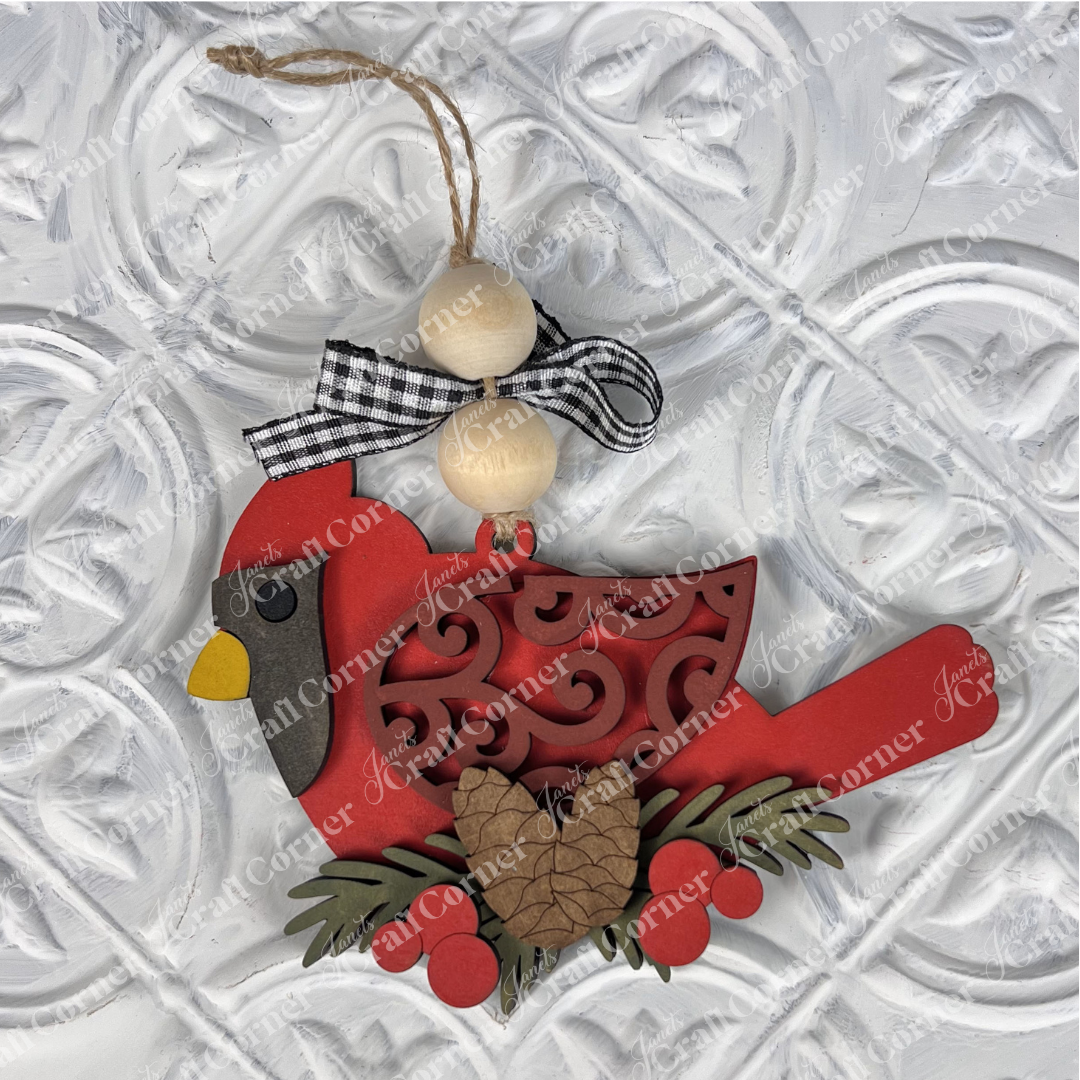 This Cardinal Ornament from Janet's Craft Corner, featured in the DIY Decoration Kits collection, is ideal for holiday décor. It showcases a yellow beak and detailed cut-out designs. Adorned with a pinecone, berries, and wooden beads topped with a ribbon, it shines elegantly against a textured white background.