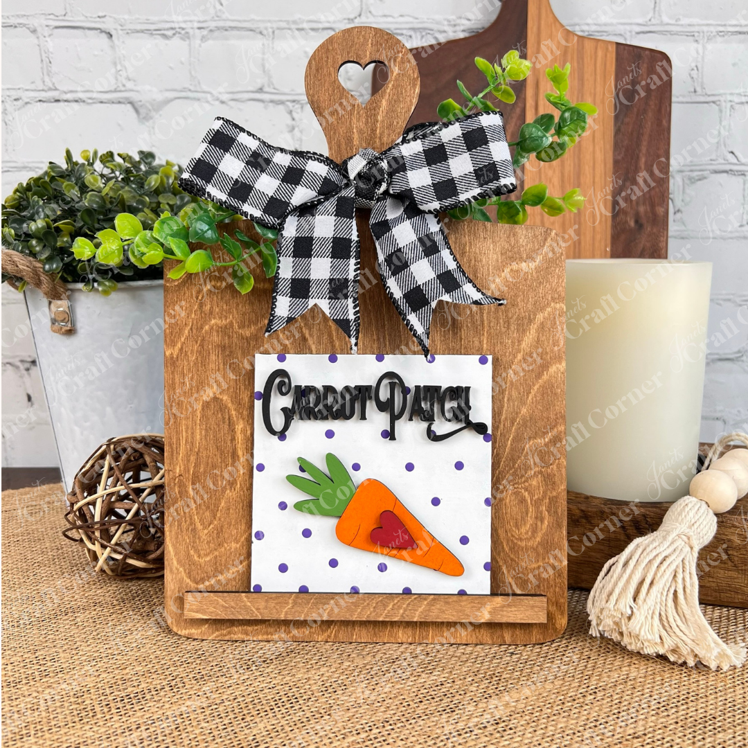Janet's Craft Corner’s Easter Bunny Squares features a wooden cutting board with a black-and-white check bow, “Carrot Patch” sign, carrot with heart, purple polka dots, plus greenery and wood accents—ideal tiered tray decor.