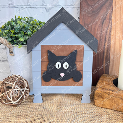 The Cat Swappable Squares by Janet's Craft Corner is a decorative gray wooden house with a black cat face, ideal for DIY seasonal decor, shown on burlap with green faux plants and wooden accents in the background.