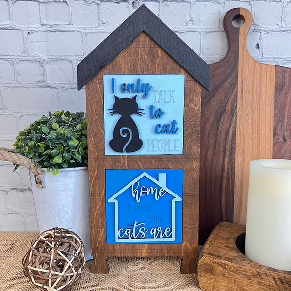Janet’s Craft Corner’s Cat Swappable Squares, a wooden house-shaped sign with cat quotes and a black cat silhouette, is perfect for DIY home decor. Styled with plants and accessories against a white brick wall for charm.