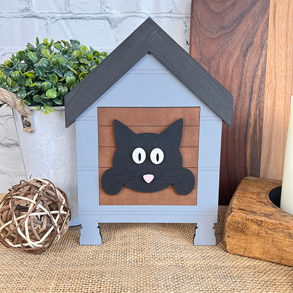 The Cat Swappable Squares by Janet's Craft Corner is a decorative gray wooden house with a black cat face, ideal for DIY seasonal decor, shown on burlap with green faux plants and wooden accents in the background.