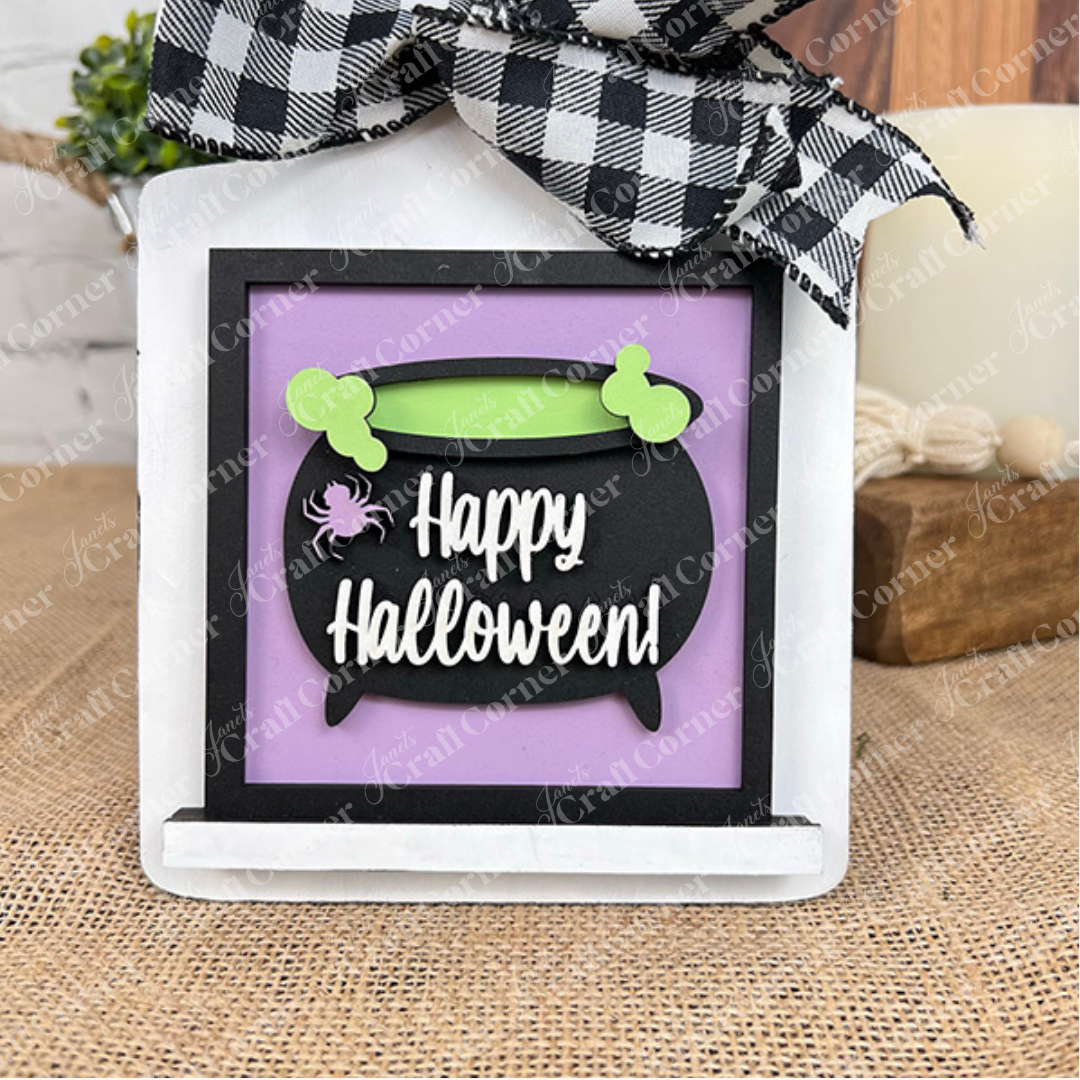 The "Oct 31 Squares" by Janet's Craft Corner features a black cauldron with green handles, a spider, and "Happy Halloween!" in white on purple—topped with a checkered bow. Festive wood cut-outs, perfect for tiered tray decor.
