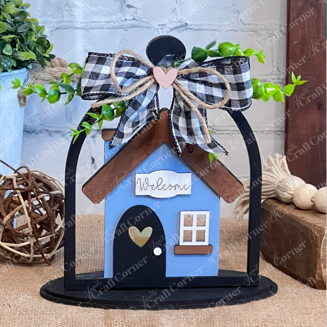 Janet's Craft Corner’s "Crafted by You!" subscription delivers a small blue house with a "Welcome" sign, black door with heart cutout, and plaid-bow arch—ideal DIY home decor for your rustic table, perfect for monthly craft kits.
