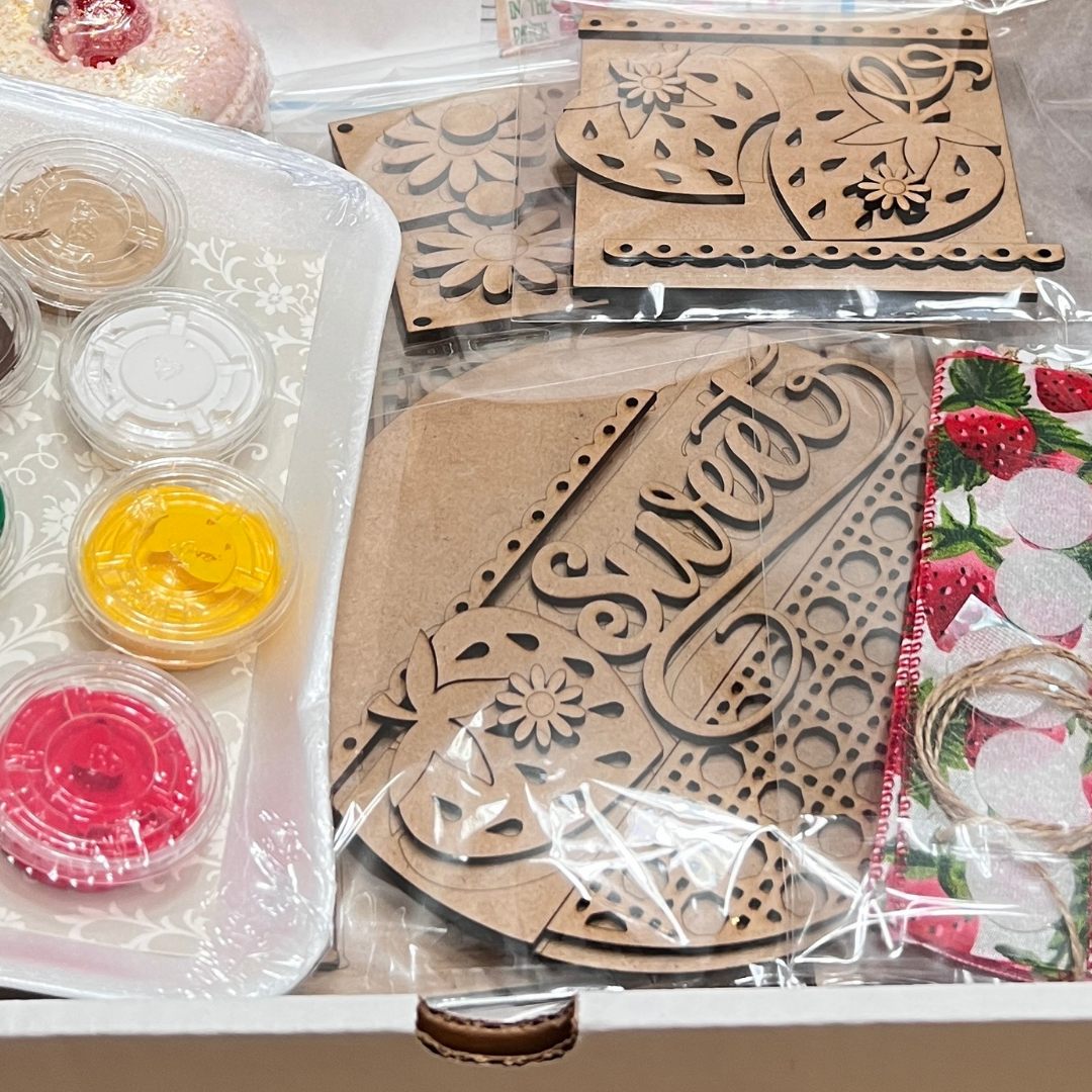 A close-up of Janet's Craft Corner's "Crafted by You!" May 2025 monthly box—a DIY home decor craft kit with wooden cutouts, colorful paints, a brush, and a strawberry napkin—perfect for creative enthusiasts.