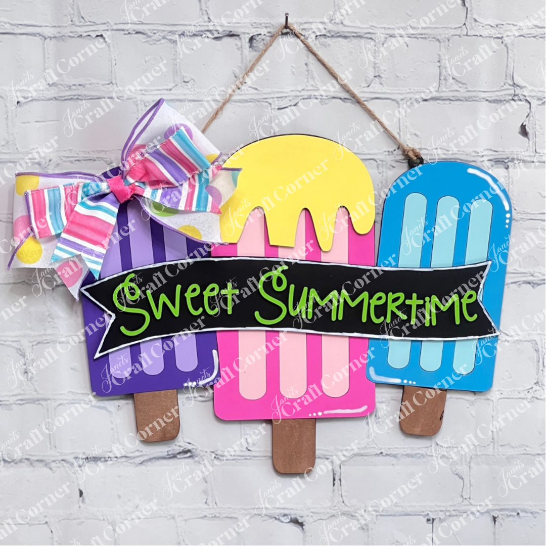 Three popsicle-shaped signs in purple, pink, and blue, made with the Janet's Craft Corner Crafted by You! monthly DIY home decor kit, hang on a white brick wall. A striped bow tops a banner reading "Sweet Summertime" in bright letters.