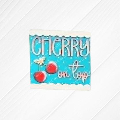 The Cherry Squares DIY seasonal home decor craft kit from Janet's Craft Corner features a blue rectangular sign with red cherries, green stems, white flowers, and "Cherry on top" in cursive—ideal for tiered trays or Swappable Squares displays.