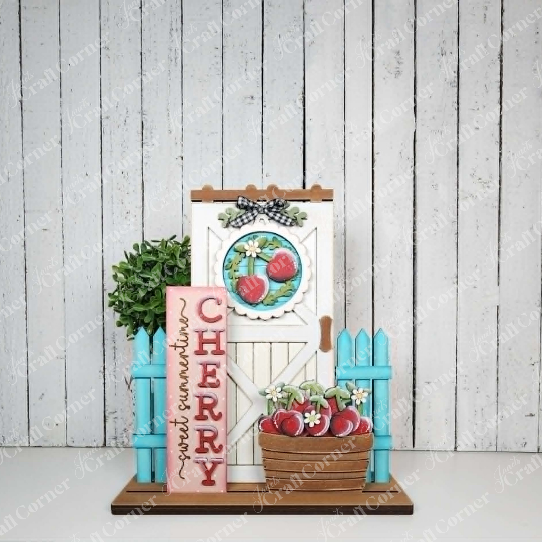 The Cherry Interchangeable Porch Set from Janet's Craft Corner is a farmhouse-style decorative wooden sign featuring a white door with cherry wreath, turquoise fence, pink “CHERRY” sign, handwritten note, potted plant, and basket of cherries.