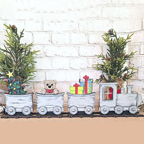 The Train Shelf Sitter with Christmas Inserts, by Janet's Craft Corner, is a festive decorative piece that includes a locomotive and two carriages. One carriage showcases a small Christmas tree decorated with ornaments and topped with a star, while the other holds a teddy bear alongside vibrant gifts. Two tiny trees positioned at the rear complete this charming holiday decor, making it ideal against a light brick wall backdrop.