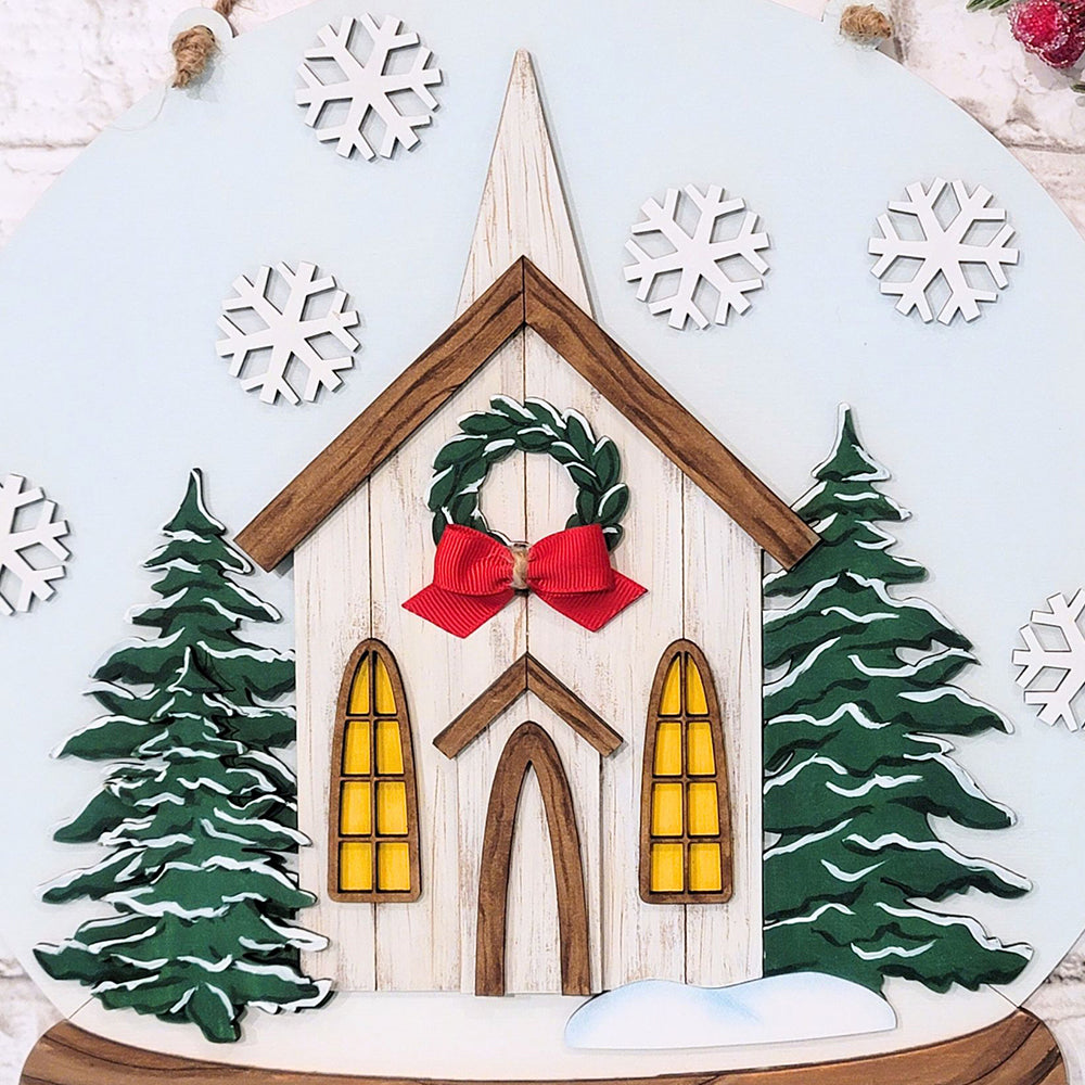 The Rustic Church Snow Globe Sign DIY Decor Kit from Janet's Craft Corner features a festive illustration of a rustic church, capturing a charming chapel adorned with a red bow wreath above its door and surrounded by snow-dusted evergreen trees. Scattered white snowflakes in the background make it perfect for home décor and creating a cozy wintery scene.